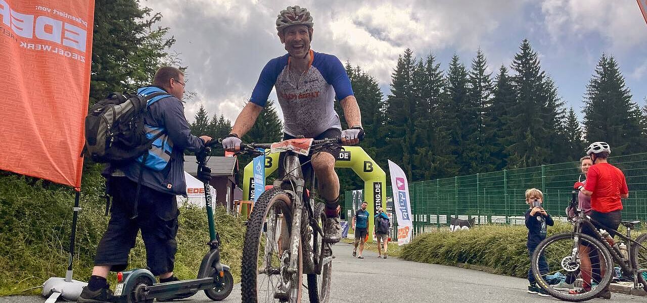 Ein mit schlamm bespritzter Mountainbiker hat gerade einen Zielbogen passiert und lächelt. 