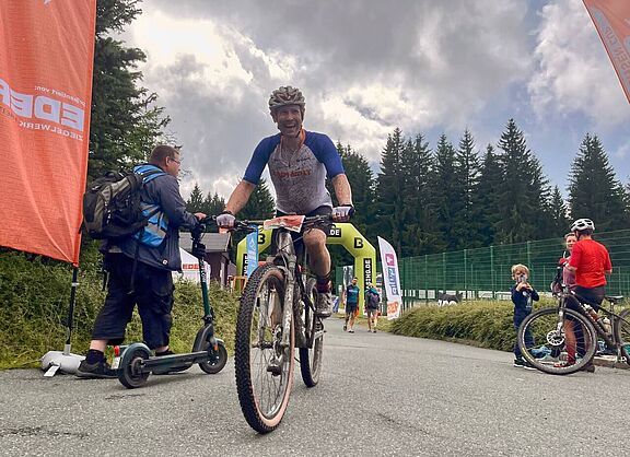 Ein mit schlamm bespritzter Mountainbiker hat gerade einen Zielbogen passiert und lächelt. 
