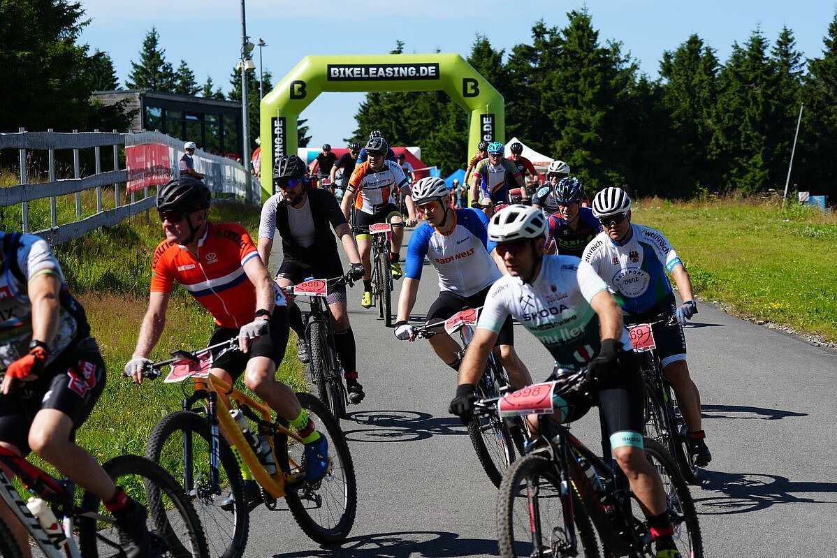 Eine Menge Mountainbiker startet zu einem Wettkampf bei Sonnenschein. Im Hintergrund ist ein Startbogen zu sehen. 