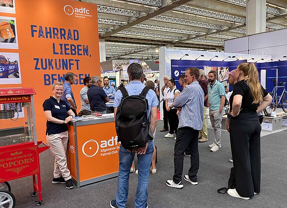 Ein Messestand des ADFC in einer großen Halle. Vor einer orangen Rückwand mit dem blauen Slogan „Fahrrad lieben, Zukunft bewegen“ steht eine lächelnde Frau im dunkelblauen ADFC-Shirt an einer Theke. Mehrere Menschen stehen davor und dahinter in Gruppen zusammen und unterhalten sich. Im Vordergrund links ist eine rote Popcornmaschine zu sehen.