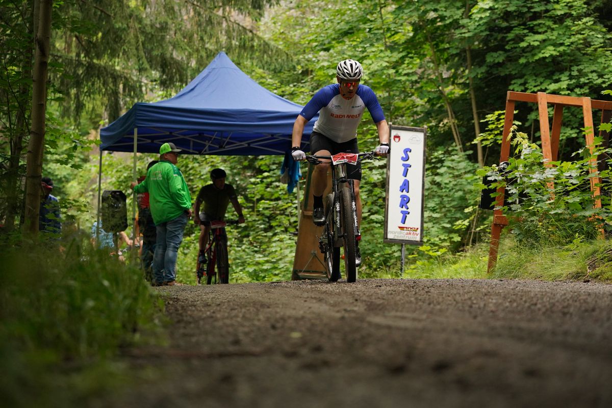 Ein Mountainbiker startet zu einem Bergzeitfahren auf einem Waldweg. Im Hintergrund ein Pavillon und ein Schild mit der Aufschrift "Start". 