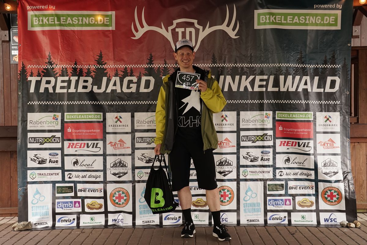 Ein Mann in Sportkleidung hält einen Starterbeutel und eine Startnummer in die Kamera. 